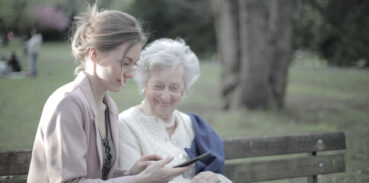 En ung kvinna och en äldre dam sitter på en parkbänk och tittar på en mobilskärm