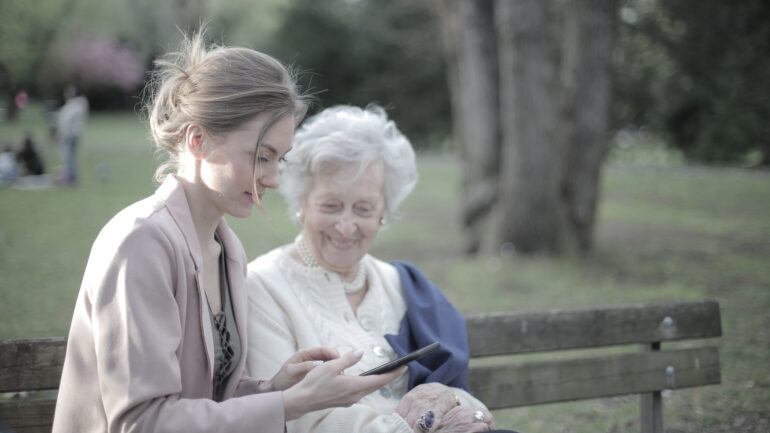 En ung kvinna och en äldre dam sitter på en parkbänk och tittar på en mobilskärm